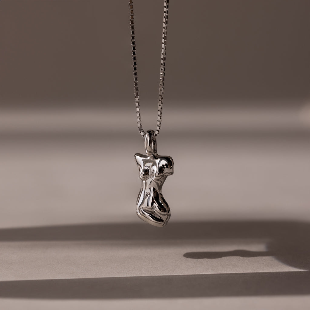 The Kinn Body Necklace in Silver features an abstract pendant on a delicate chain, casting a soft shadow—an elegant and empowering accessory inspired by the Venus de Milo.