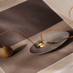 The Duo Pebble Necklace in 18K Gold, featuring a sculptural pendant, is shown on a magazine page with a plate, fork, and pear—ideal for minimal jewelry lovers.