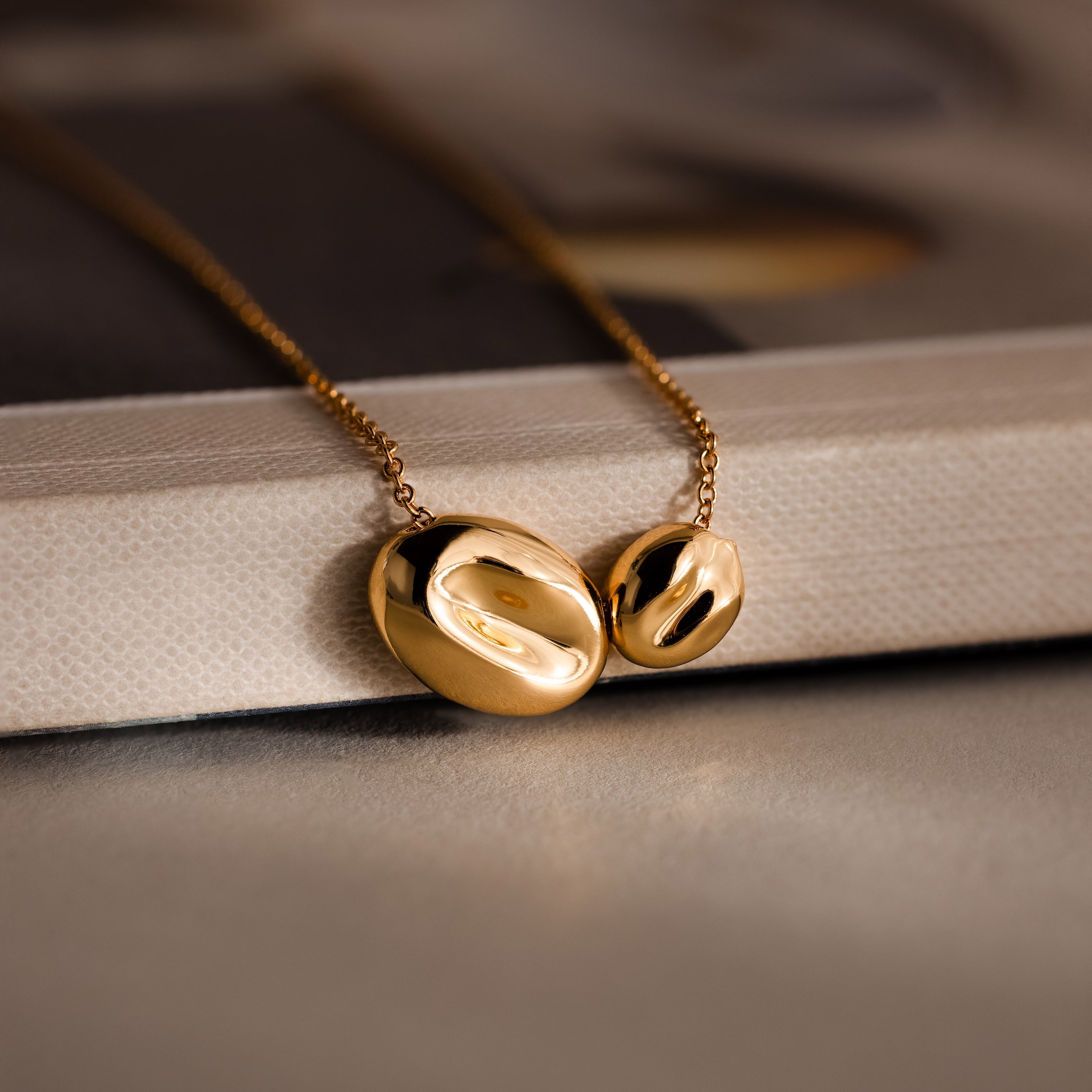The Duo Pebble Necklace features two smooth, oval gold pendants displayed on a beige textured surface.