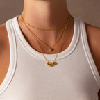 A person in a white tank top wears minimal jewelry: three layered gold necklaces, including a sculptural pendant and the Duo Pebble Necklace in 18K Gold, each with a unique design.