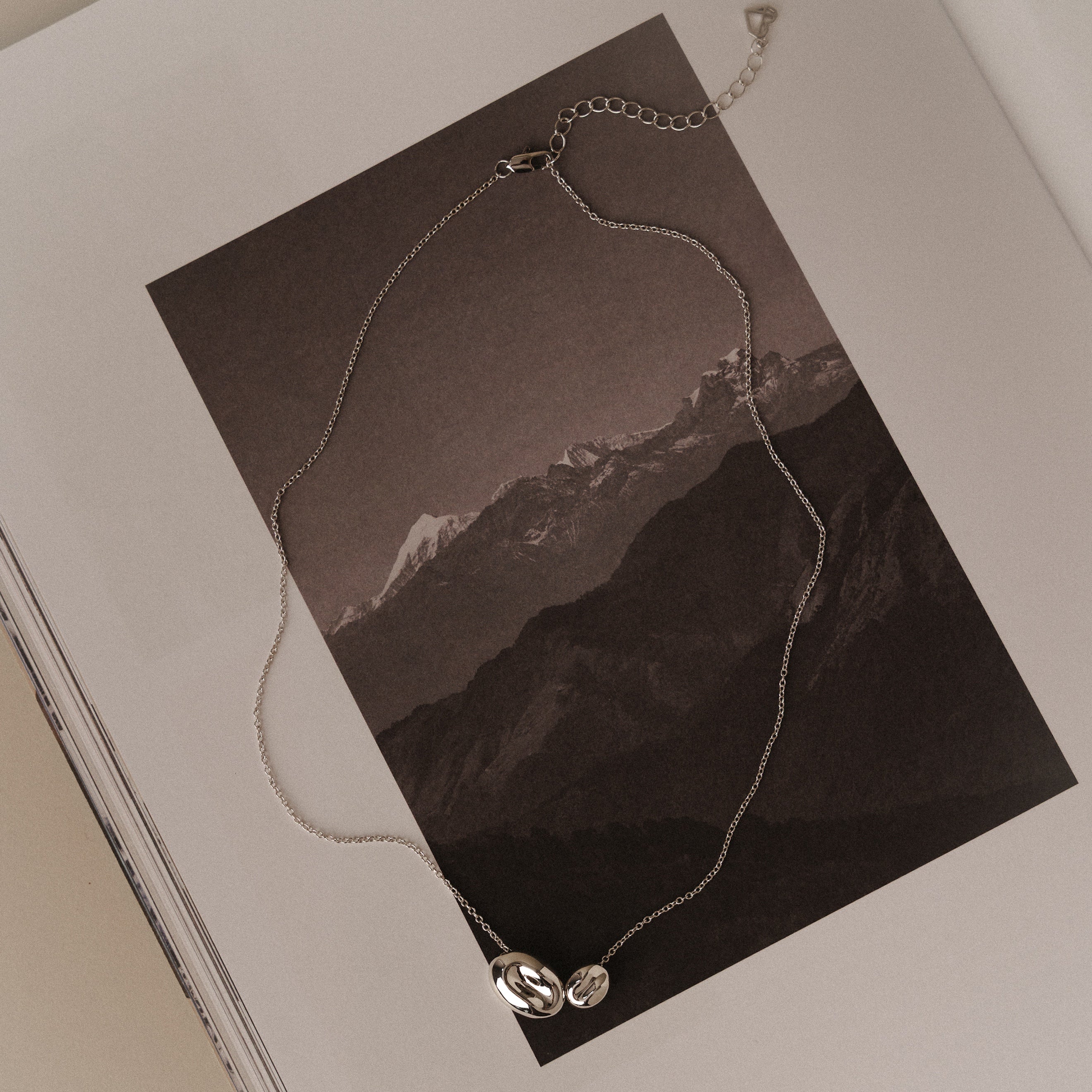 The Duo Pebble Necklace in Silver, featuring two dainty pendants, rests on an open book with a black and white mountain photo, lending subtle elegance to the scene.