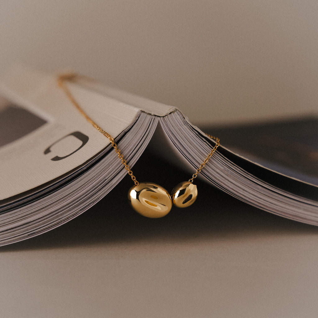 The Duo Pebble Necklace in 18K Gold—a sculptural pendant—draped over an open magazine, styled minimally with soft neutral lighting and a clean background.