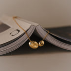 The Duo Pebble Necklace in 18K Gold—a sculptural pendant—draped over an open magazine, styled minimally with soft neutral lighting and a clean background.