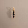 The Ombre Lace Onyx Charm is a gold pendant featuring five small black stones in a vertical line, casting an elegant shadow on a beige surface.
