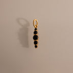 The Ombre Lace Onyx Charm is a gold pendant featuring five small black stones in a vertical line, casting an elegant shadow on a beige surface.