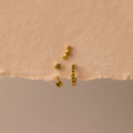 The Peridot Earring Trio Set, featuring four gold pieces with green stones including a pavé huggie hoop, is displayed on textured, torn beige paper.