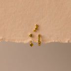 The Peridot Earring Trio Set, featuring four gold pieces with green stones including a pavé huggie hoop, is displayed on textured, torn beige paper.
