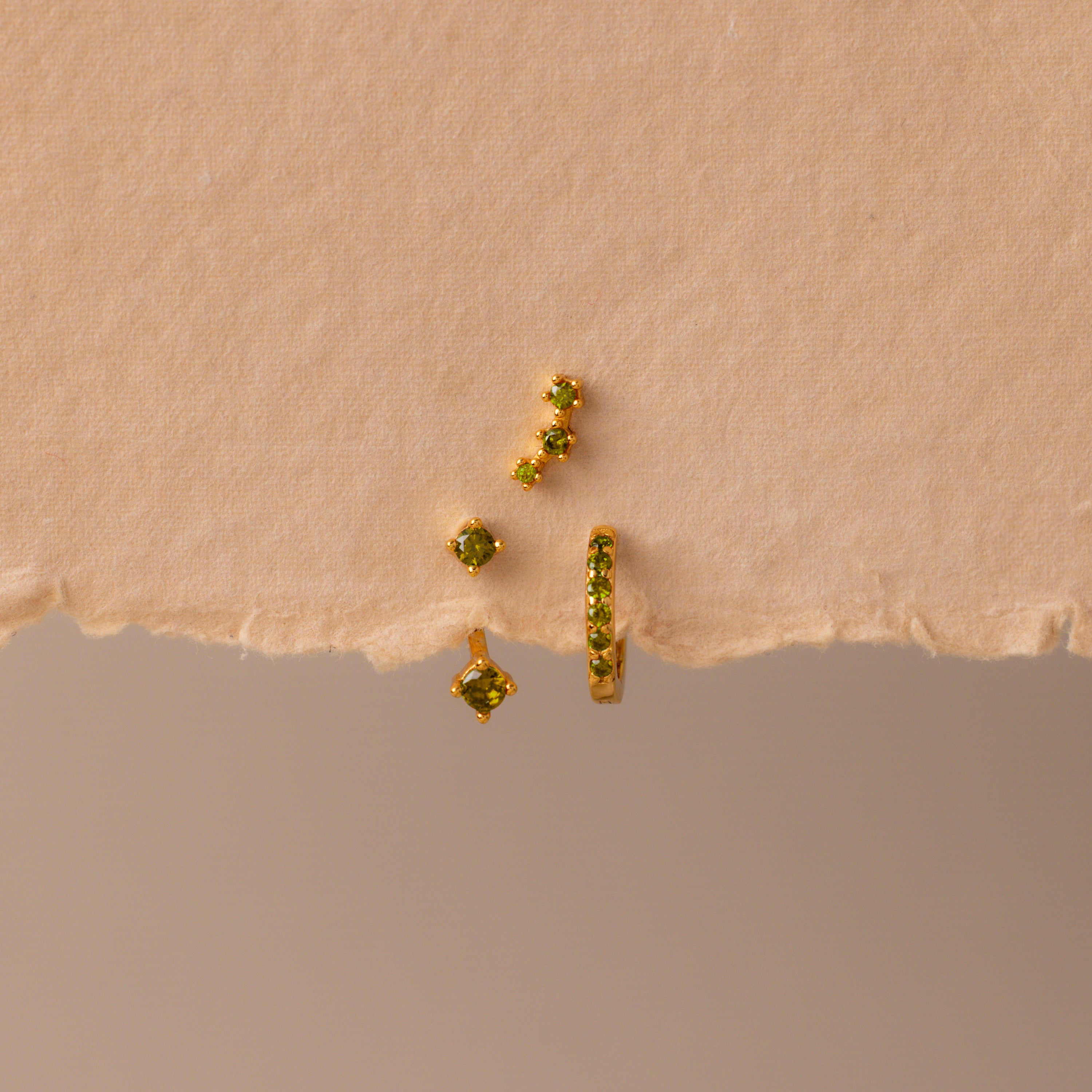 The Peridot Earring Trio Set, featuring four gold pieces with green stones including a pavé huggie hoop, is displayed on textured, torn beige paper.