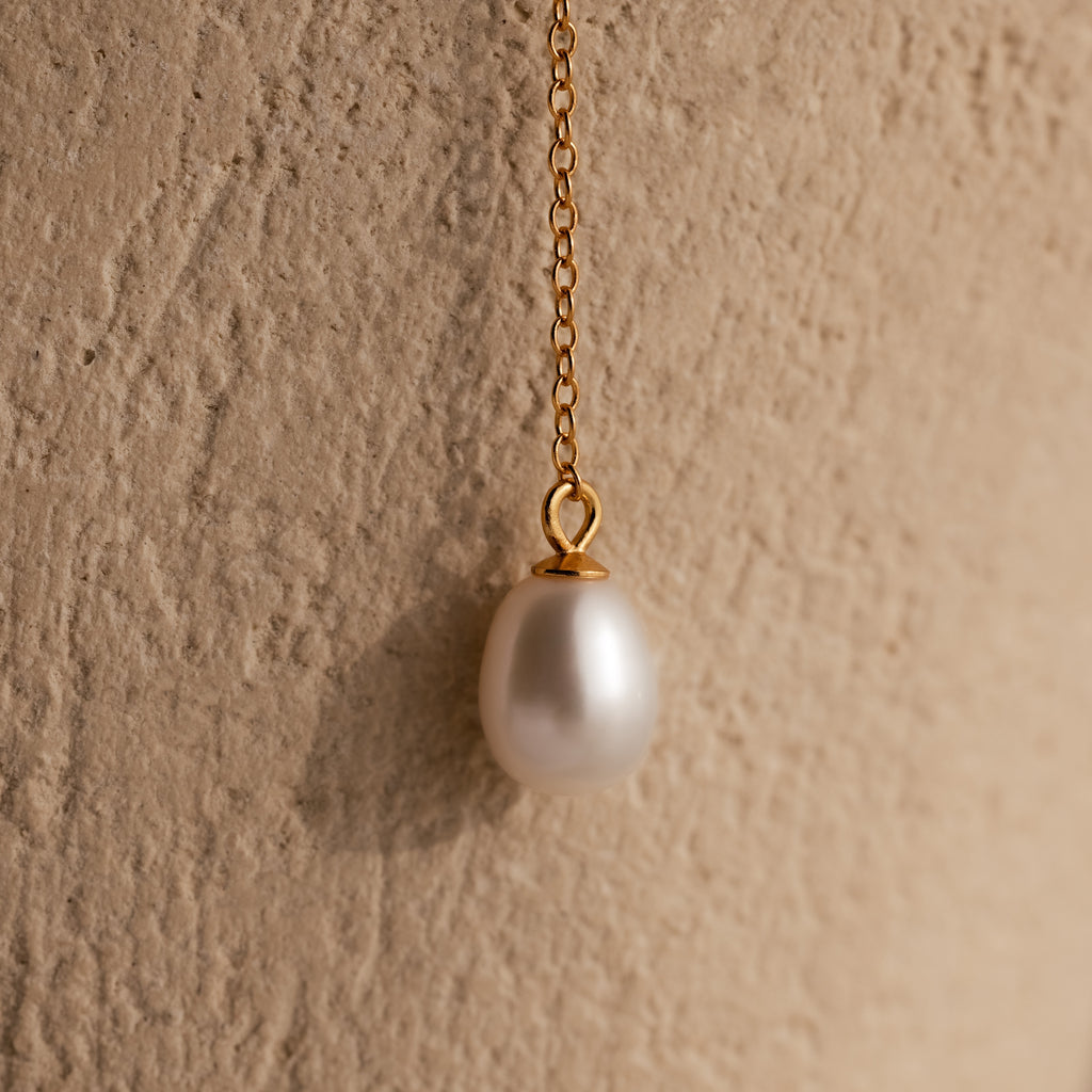 The Pearl Drop Lariat Necklace in 18K Gold features a delicate vertical chain with a single white pearl pendant, elegantly displayed against a textured beige background.