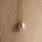 The Pearl Drop Lariat Necklace in 18K Gold features a delicate vertical chain with a single white pearl pendant, elegantly displayed against a textured beige background.