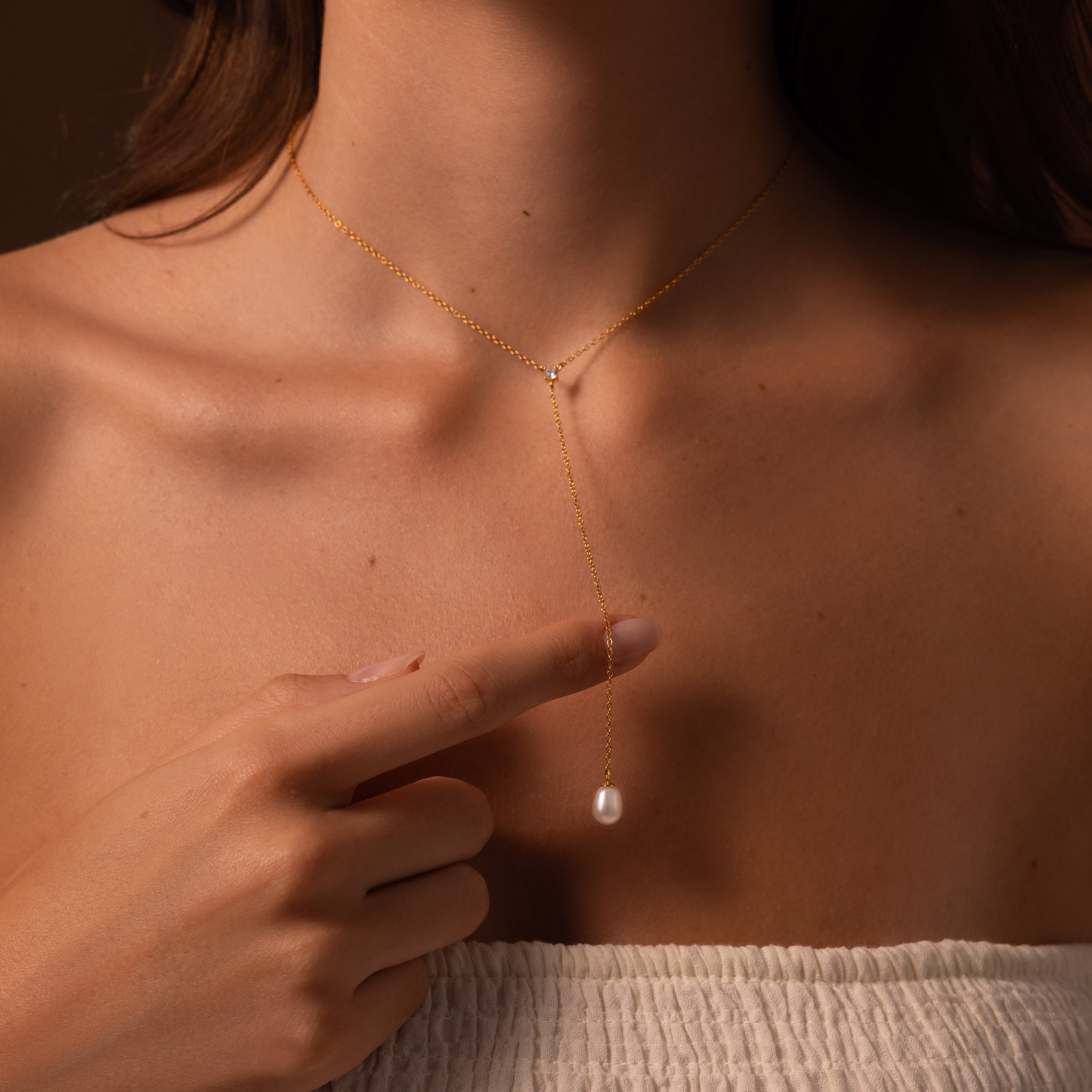 A woman in a strapless top points to the pendant of the elegant Pearl Drop Lariat Necklace, featuring a delicate gold Y-shaped chain and a beautiful pearl drop.