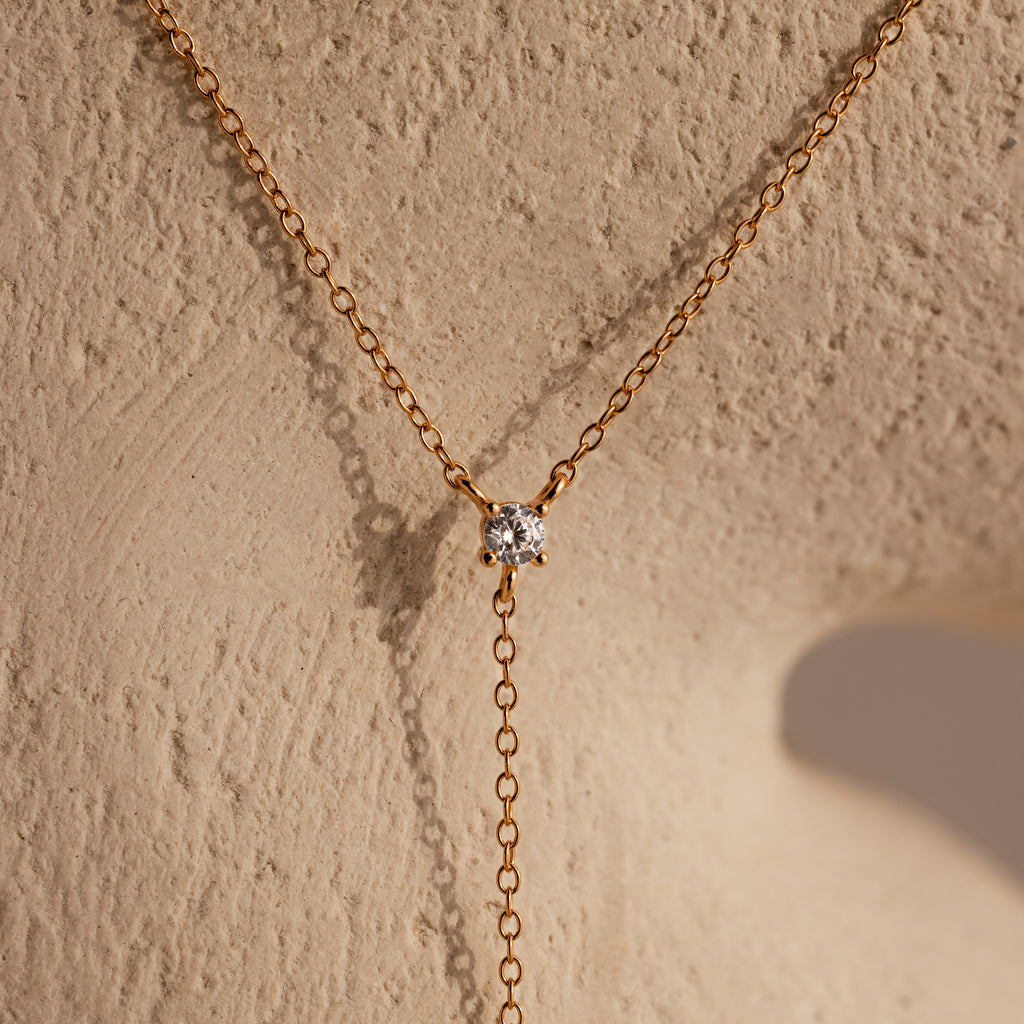 The Pearl Drop Lariat Necklace in 18K Gold, with its elegant design, adds a refined touch to any outfit, similar to a gold chain necklace with a round diamond pendant displayed on a textured beige surface.