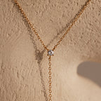 The Pearl Drop Lariat Necklace in 18K Gold, with its elegant design, adds a refined touch to any outfit, similar to a gold chain necklace with a round diamond pendant displayed on a textured beige surface.