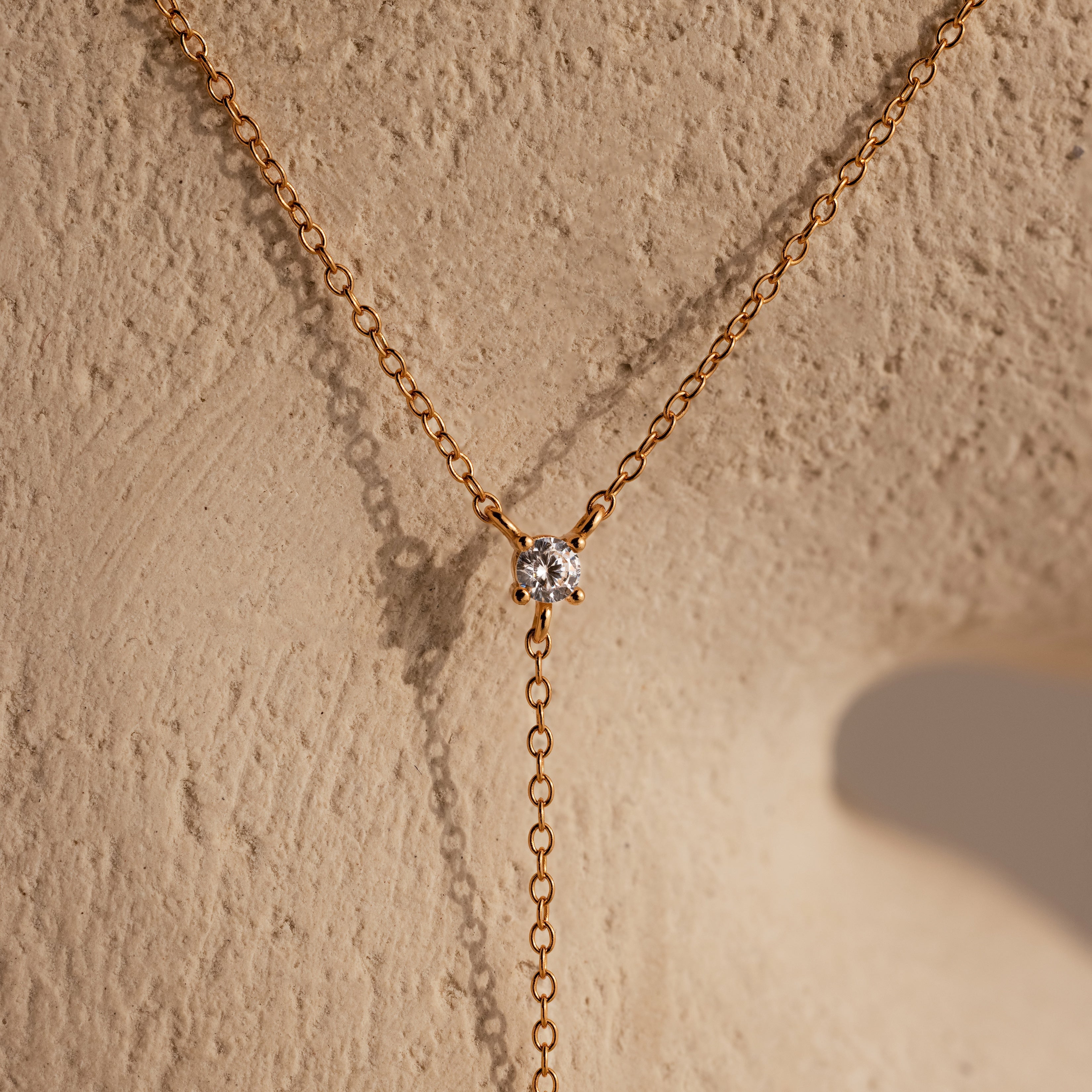 The Pearl Drop Lariat Necklace in 18K Gold, with its elegant design, adds a refined touch to any outfit, similar to a gold chain necklace with a round diamond pendant displayed on a textured beige surface.