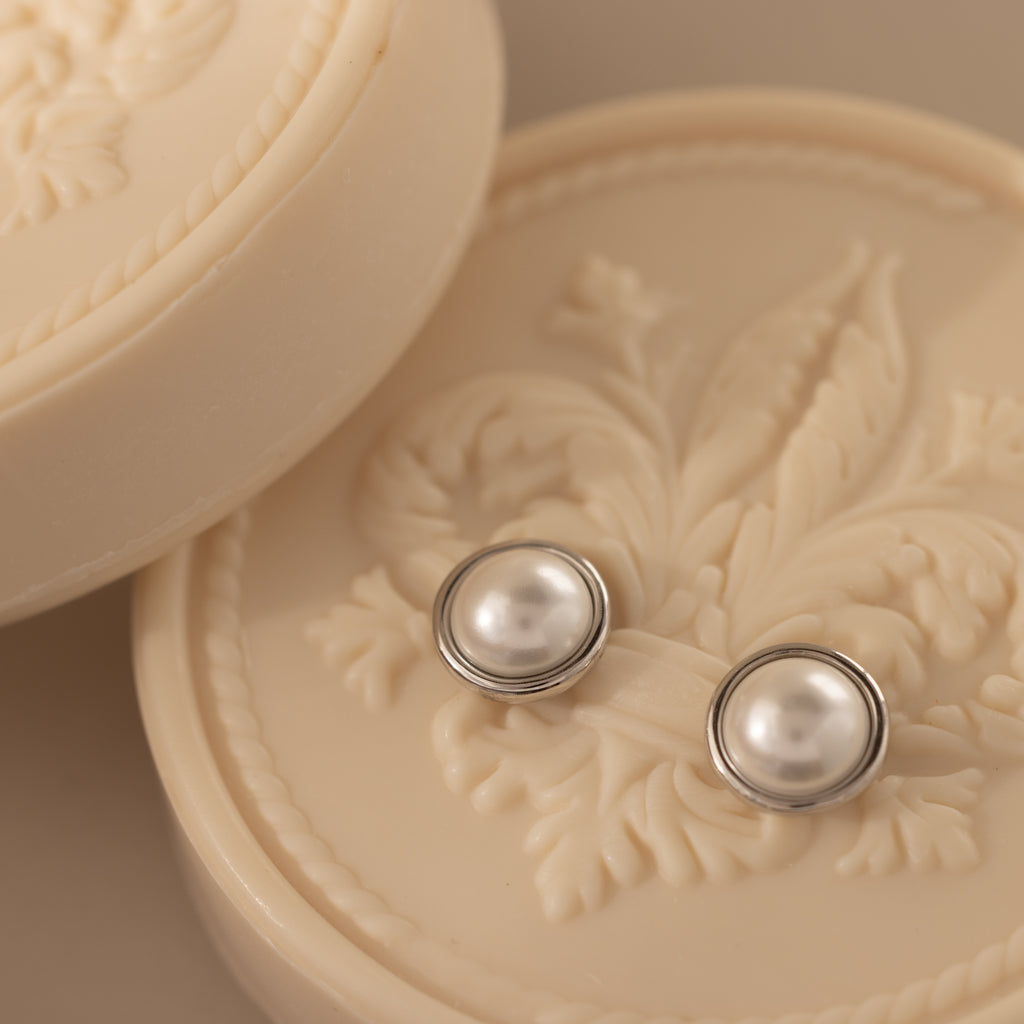 The Pearl Bezel Studs in Sterling Silver are displayed elegantly atop an ornate, cream-colored, embossed soap container.