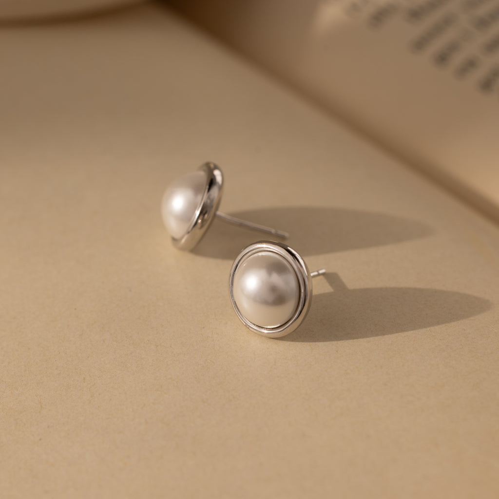 The Pearl Bezel Studs, featuring silver rims, rest on a beige surface, their elegant luster showcasing the timeless beauty of classic jewelry, with a blurred book in the background.