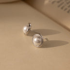 The Pearl Bezel Studs, featuring silver rims, rest on a beige surface, their elegant luster showcasing the timeless beauty of classic jewelry, with a blurred book in the background.
