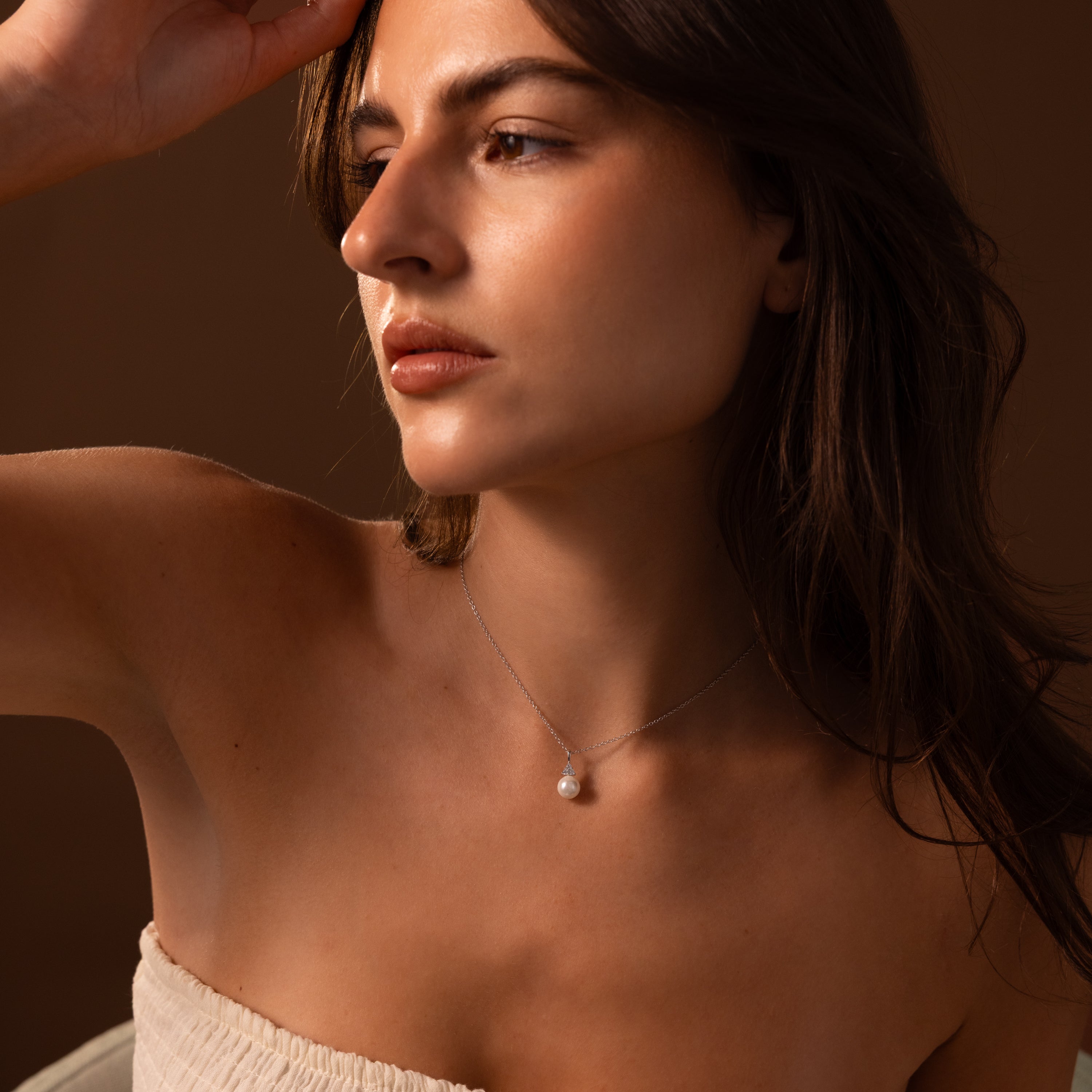 A woman in a strapless top gazes sideways, highlighting the Diamond Halo Pearl Necklace in Sterling Silver—a timeless piece—set against a brown background.