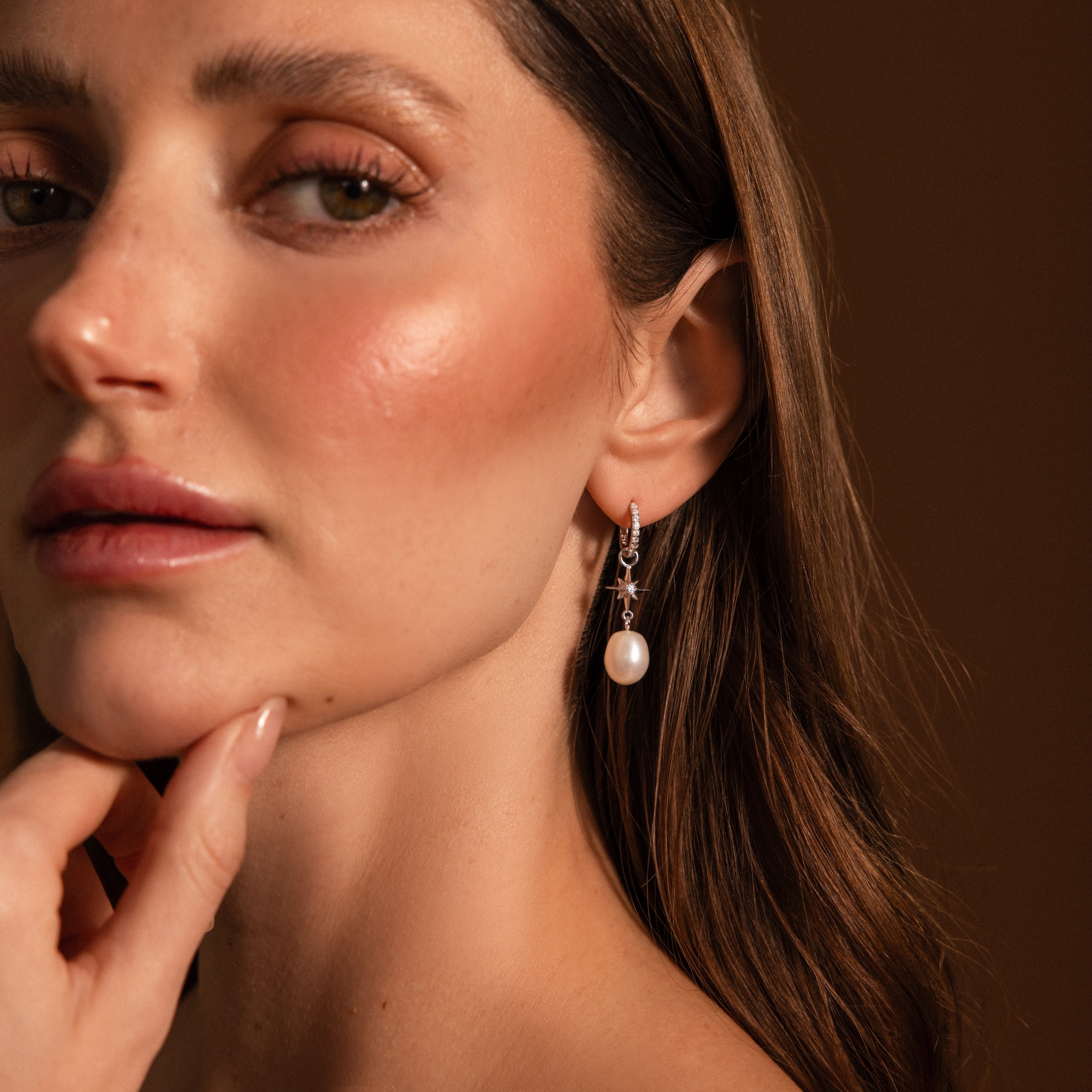 Woman with glowing skin wears North Star Pearl Hoops in Sterling Silver, softly gazing and touching her chin as the shimmering star detail stands out against a brown background.