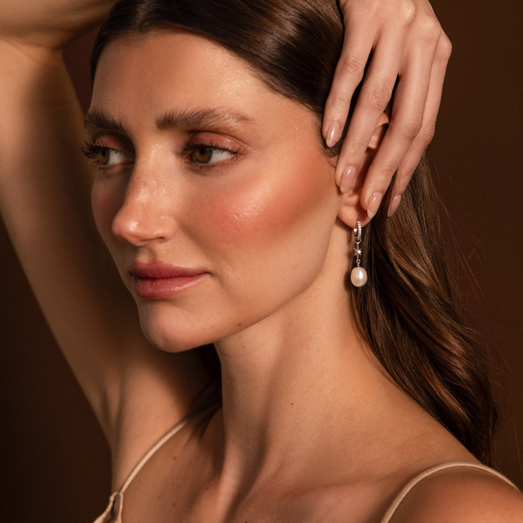 Woman with wavy brown hair wears North Star Pearl Hoops in Sterling Silver, touching her head with one hand.