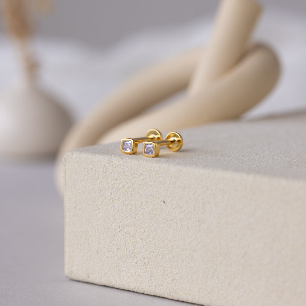 A pair of Square Bezel Birthstone Flatback Studs in Alexandrite (June), featuring small gold square CZ gemstones, rests on a textured beige surface.