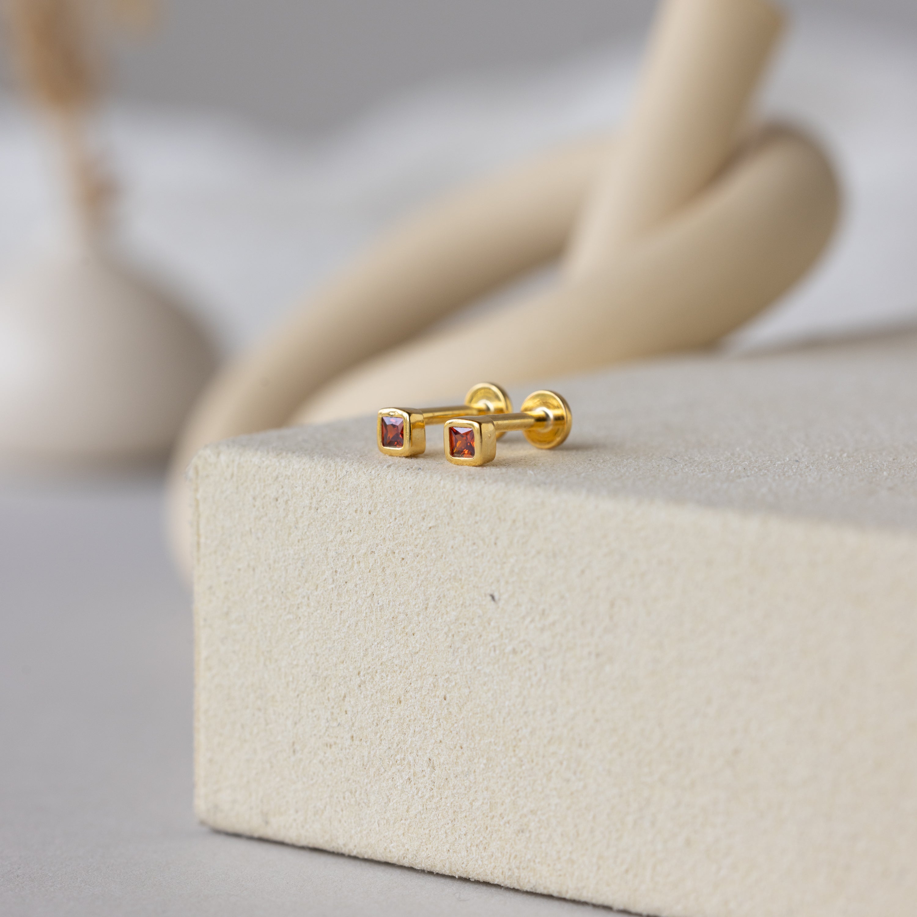 A pair of Square Bezel Birthstone Flatback Studs in Garnet (January), shown on a textured beige surface.