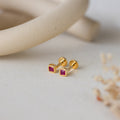 Square Bezel Ruby Flatback Studs, featuring small CZ gemstones set in a gold square bezel, are displayed on a light, textured surface with soft decor.