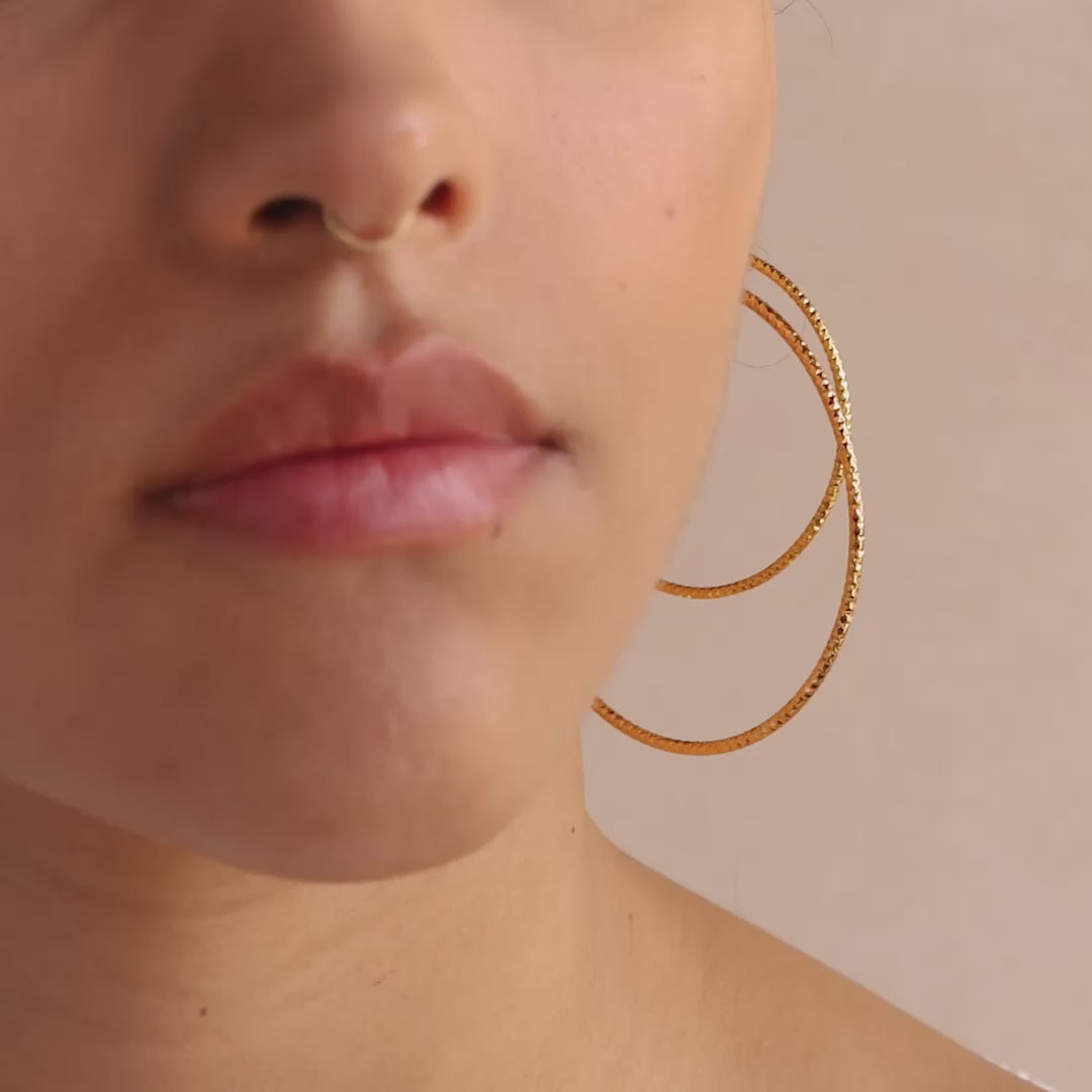 Model showcasing two large, textured gold hoop earrings. The hammered finish on the hoops reflects light, creating a shimmering effect.