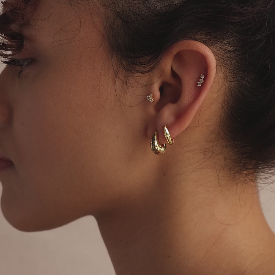 Close-up of a model wearing two gold huggie earrings—one thick and oval-shaped, the other sleek and leaf-shaped—styled with dainty pavé studs.