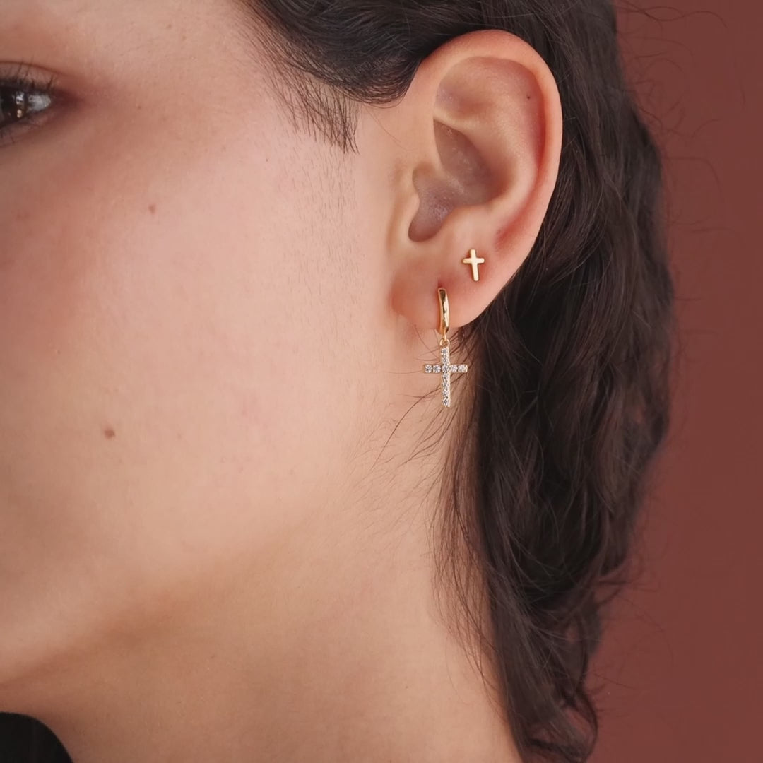 Model showcasing gold huggie hoop earrings with dangling pave cross charms, stacked with dainty cross stud earrings.