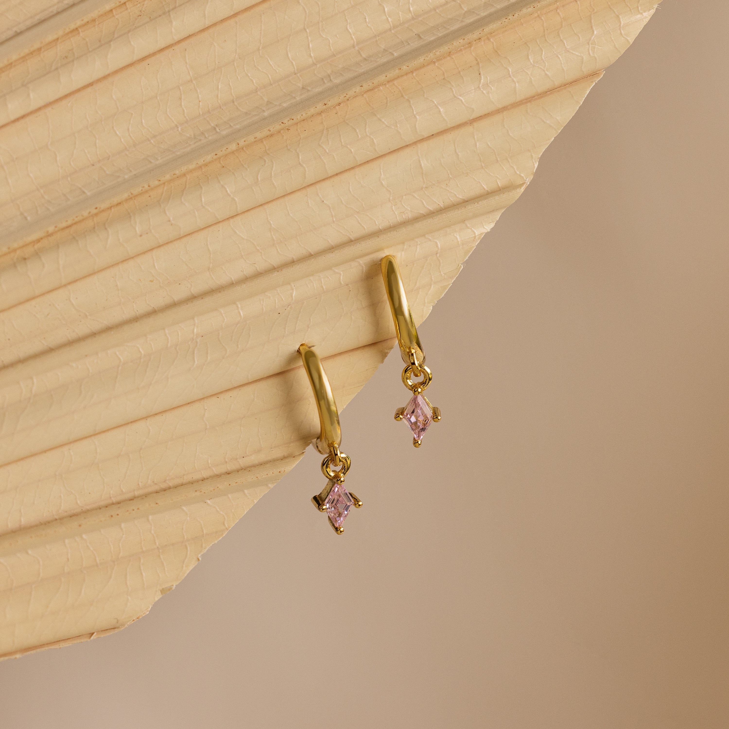 Ruby Drop Huggies, dainty gold hoop earrings with small pink gemstone charms, are displayed on a beige textured surface.