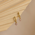 Pink Tourmaline Drop Huggies with small, pink, diamond-shaped pendants are displayed on a textured beige surface.