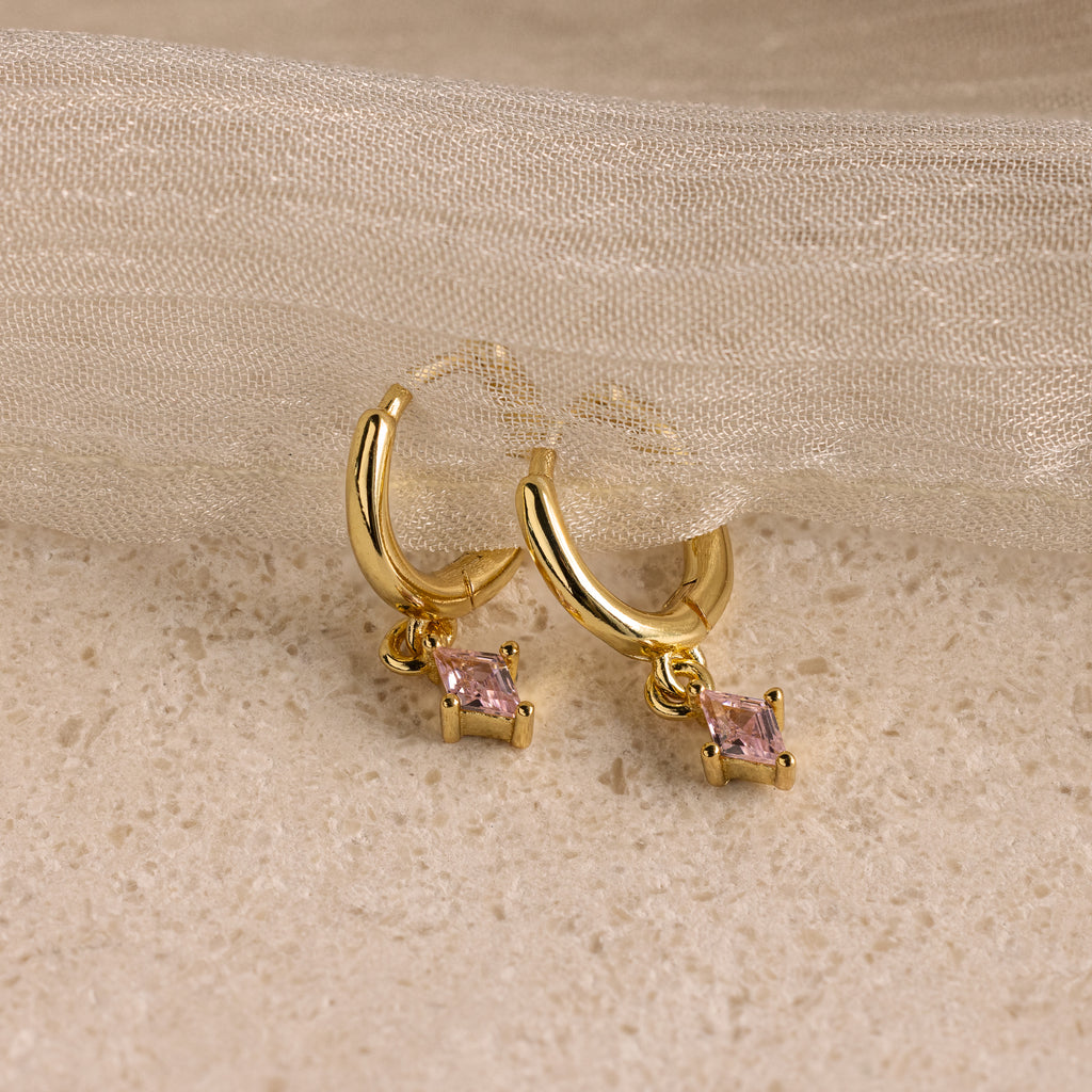 Pink Tourmaline Drop Huggies, shown on a textured beige surface with sheer fabric—ideal as personalized jewelry or birthstone earrings featuring small pink gemstones.