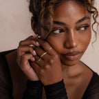 Woman resting her face on her clasped hands with a variety of gold green peridot rings on a neutral background