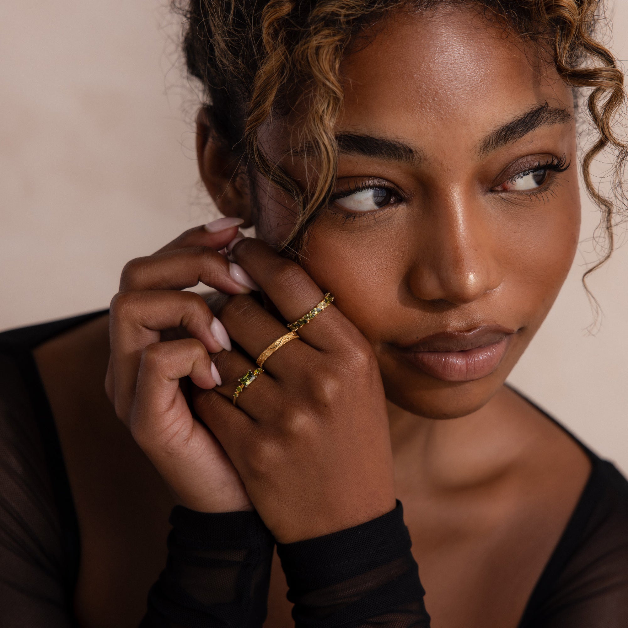 Woman resting her face on her clasped hands with a variety of gold green peridot rings on a neutral background