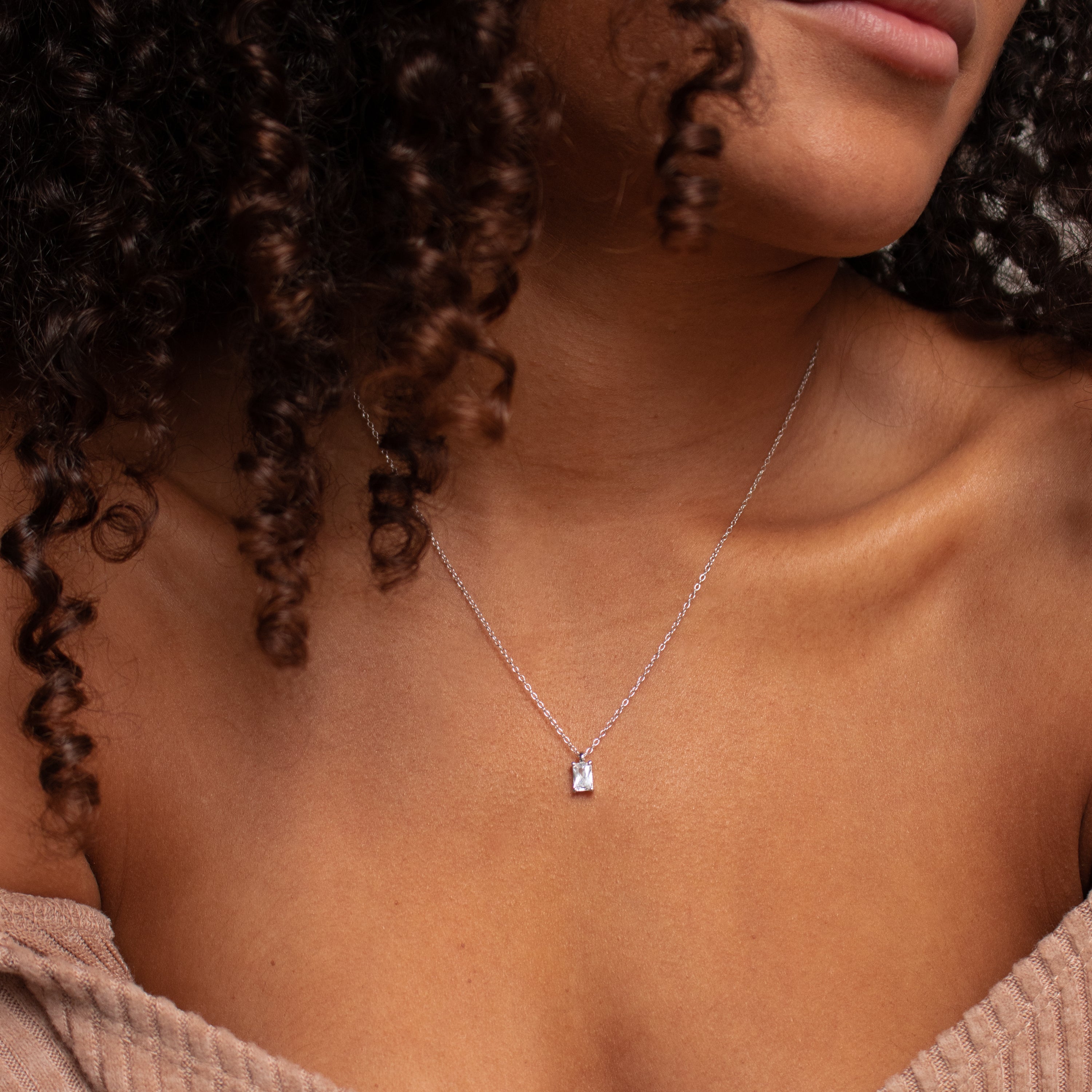A woman with curly hair wears the Sage Diamond Necklace—a luxury silver chain with a small, rectangular pendant—gracefully displayed on her bare neck.
