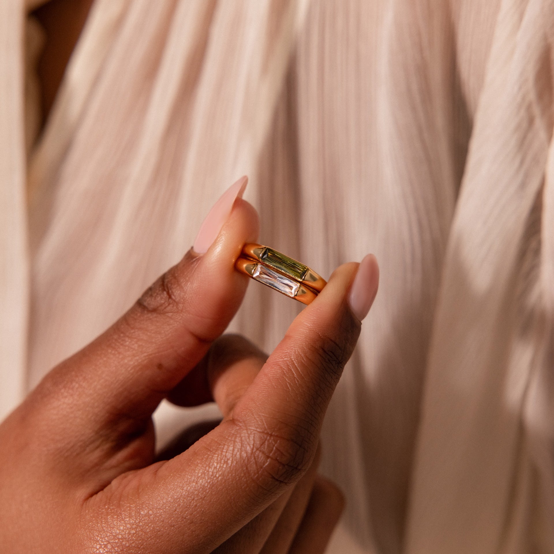 A hand with nude nails displays the Baguette Signet Ring in gold with a rectangular gemstone, set against a cream fabric backdrop.