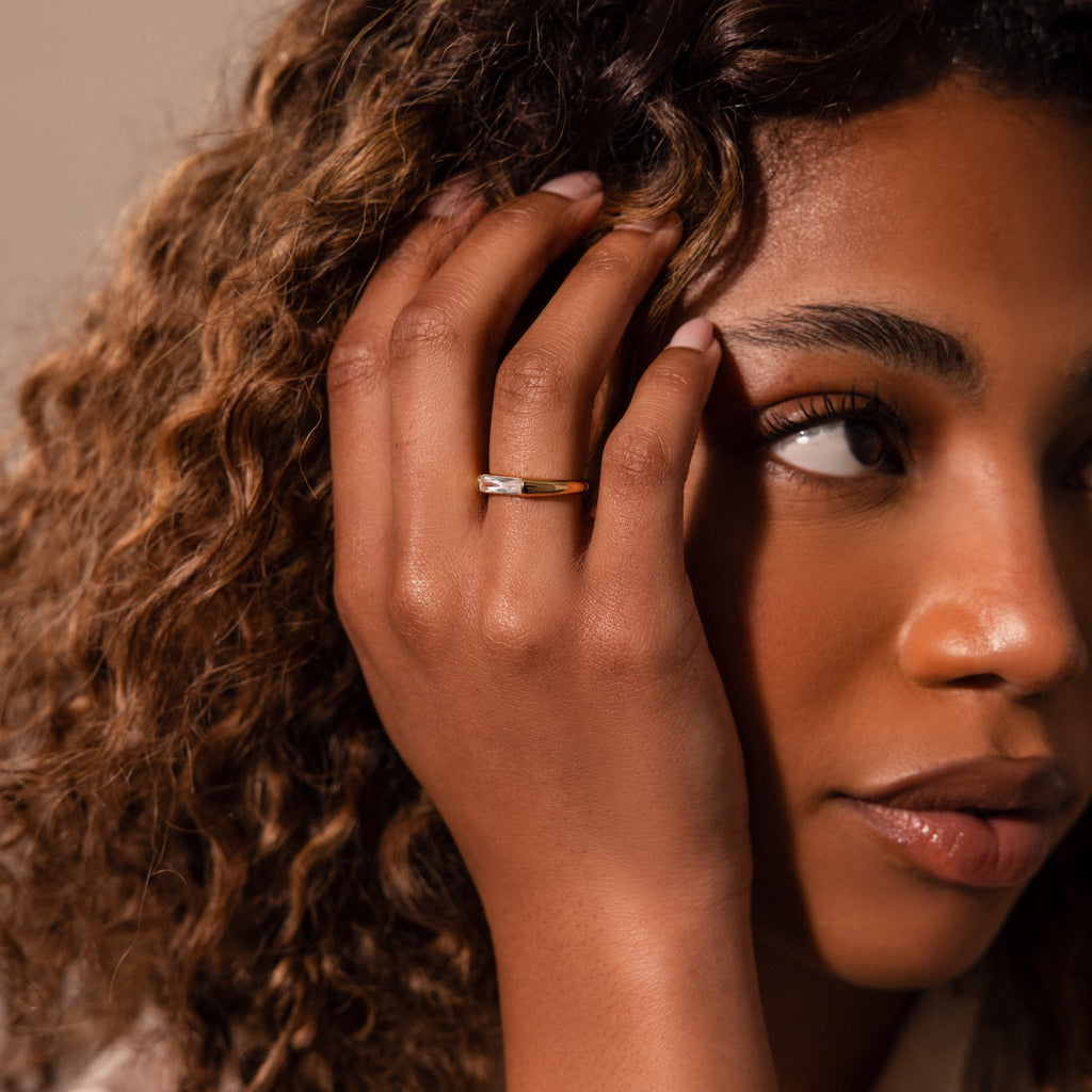 A woman with curly hair touches her face, wearing the Diamond Baguette Signet Ring—a modern gold signet featuring a small diamond—on her ring finger.