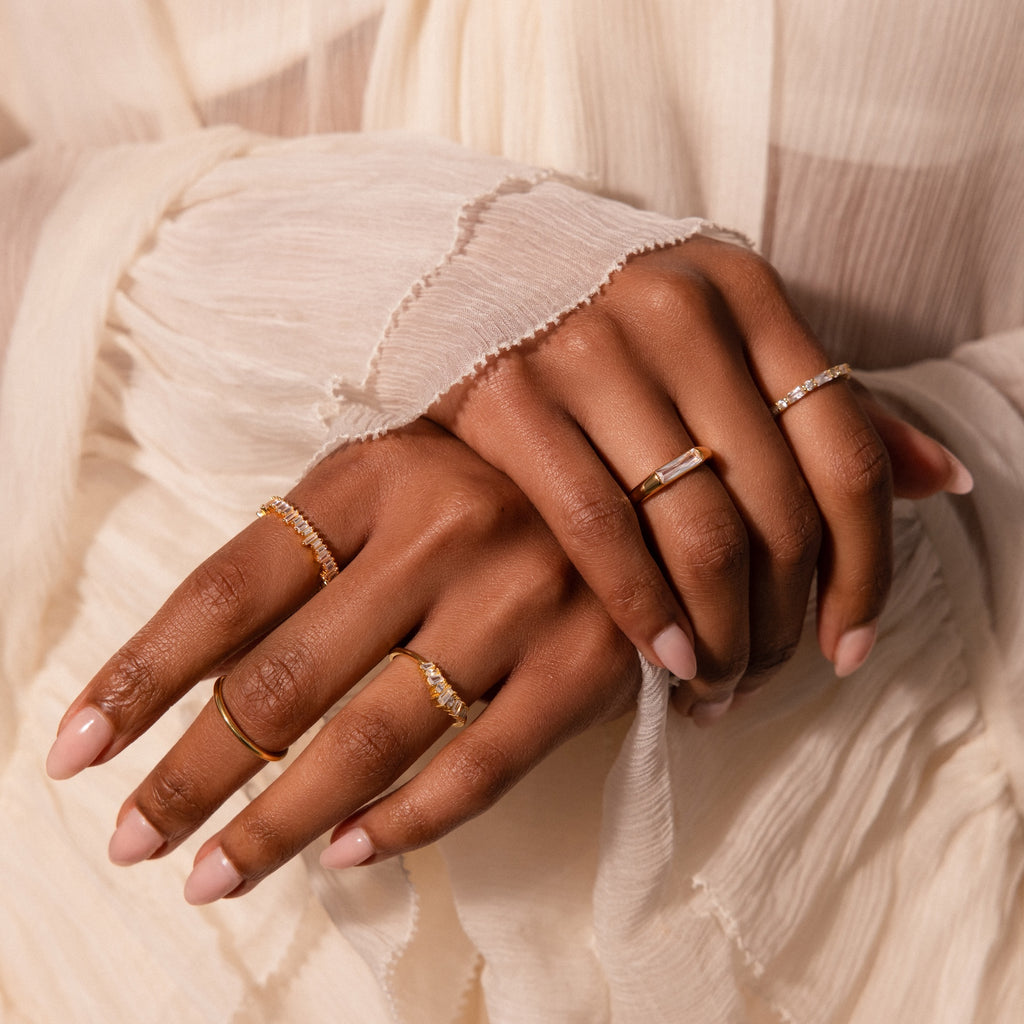 Hands with nude nails showcasing delicate gold and diamond rings, featuring the Baguette Signet Ring, against a cream pleated fabric background.