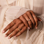 Hands with nude nails showcasing delicate gold and diamond rings, featuring the Baguette Signet Ring, against a cream pleated fabric background.