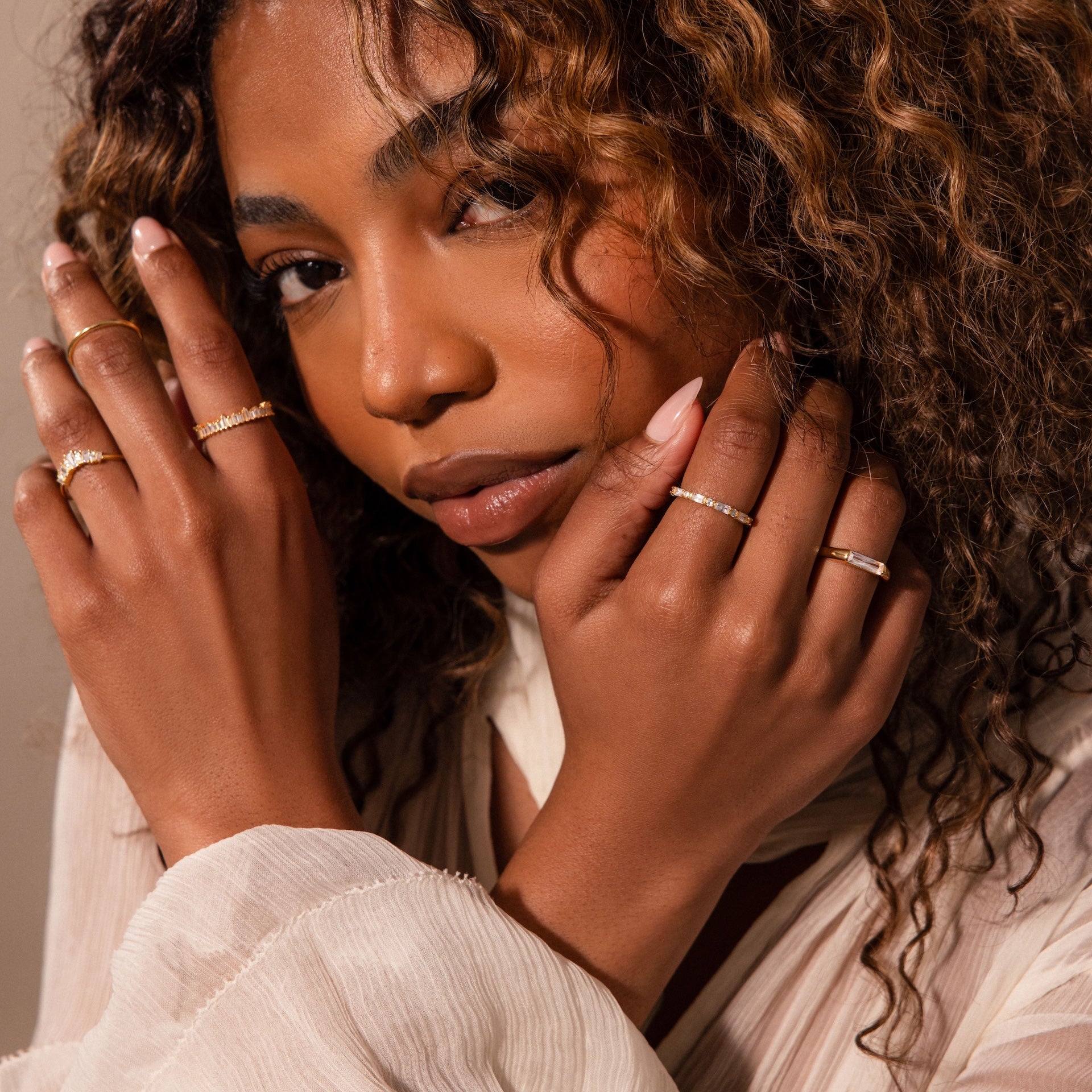Woman with curly hair wears the Diamond Baguette Signet Ring with gold bands and a light, textured blouse, posing with hands near her face.