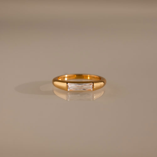 Diamond Baguette Signet Ring: This modern gold ring features a rectangular diamond centerpiece, showcased on a reflective beige surface.