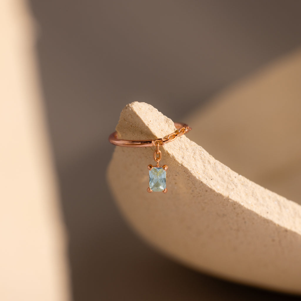 The Aquamarine Drop Chain Ring, featuring a delicate gold chain and a small emerald-cut light blue gemstone charm, rests on a textured beige surface.