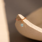 The Aquamarine Drop Chain Ring, featuring a delicate gold chain and a small emerald-cut light blue gemstone charm, rests on a textured beige surface.