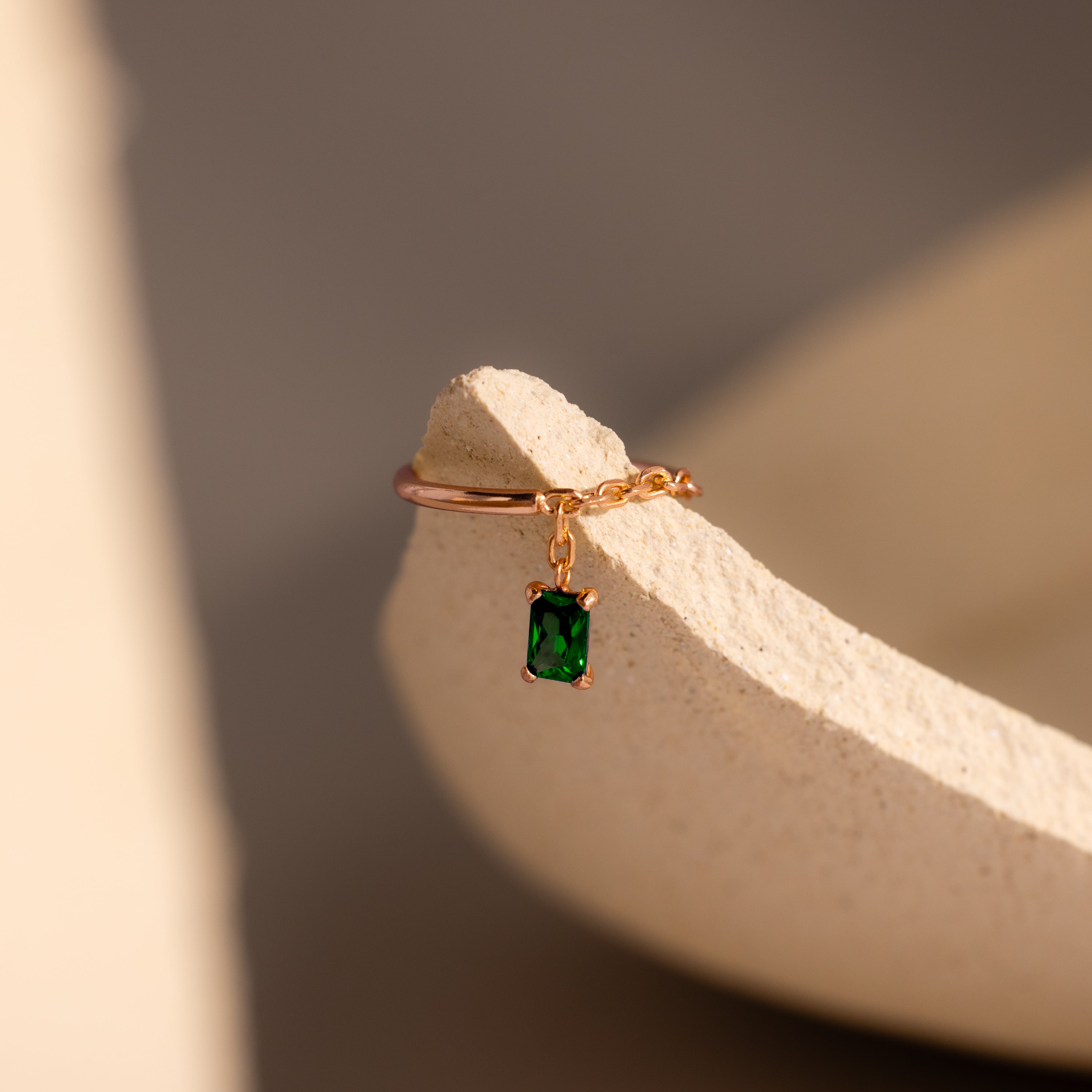 The Emerald Drop Chain Ring features a gold chain band with a dangling emerald-cut green stone, showcased on a textured beige surface.