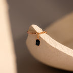 Onyx Drop Chain Ring featuring a small emerald-cut black gemstone charm, displayed on a textured beige surface.
