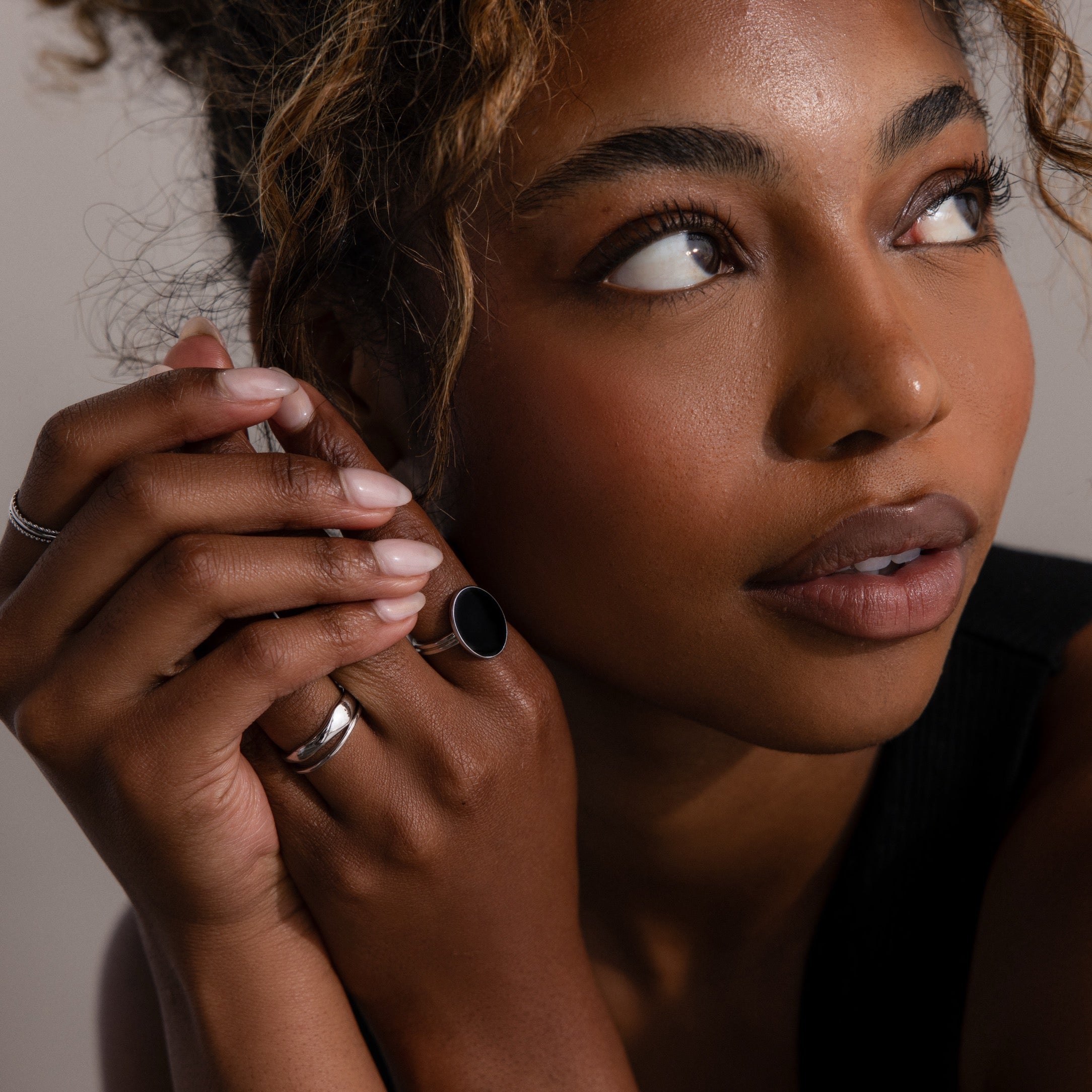 Woman with curly hair looks upward, hands near face adorned with sculptural jewelry, wearing the Duo Form Ring Set in Silver and natural makeup.