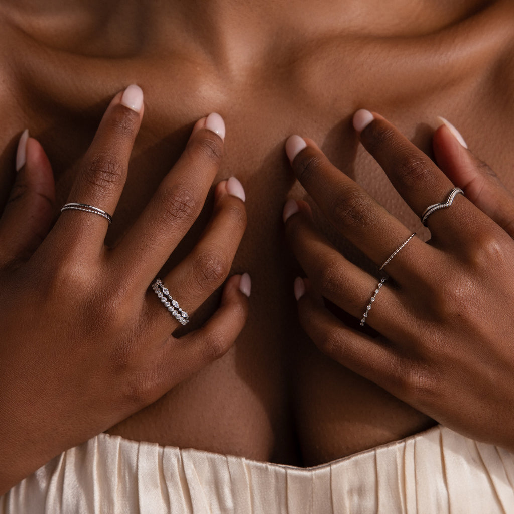 Close-up of elegant hands with manicured nails wearing the Crystal Stacking Ring Set in Silver, paired with various other rings, resting on a womans chest in a cream dress.