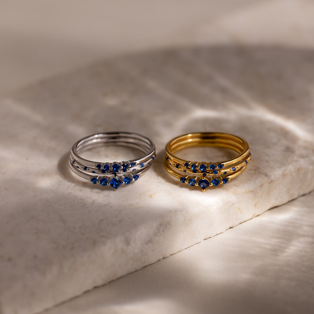 Two sets of stacking rings, three silver and three gold, with blue stones on a marble surface.