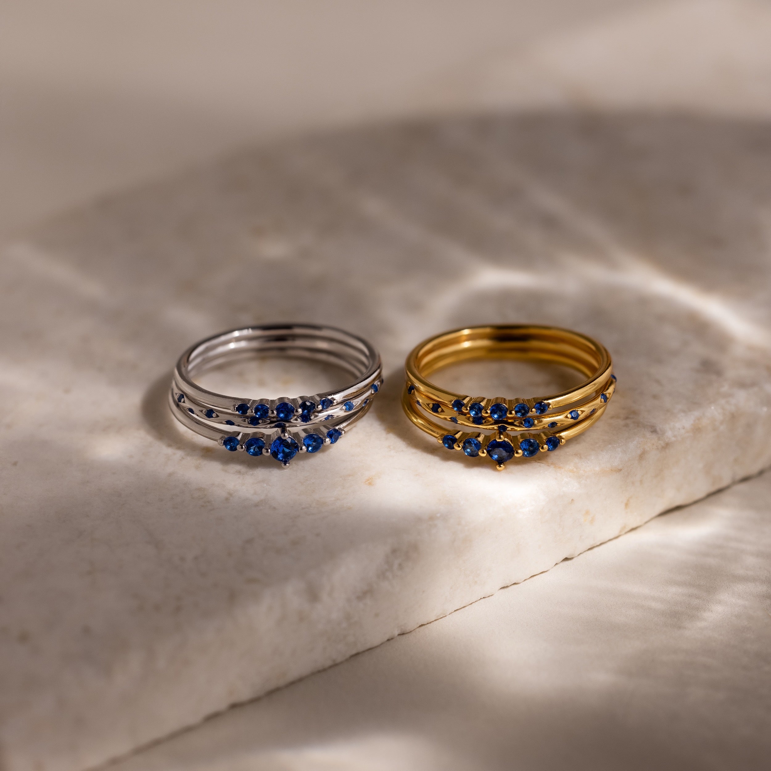 Two sets of stacking rings, three silver and three gold, with blue stones on a marble surface.